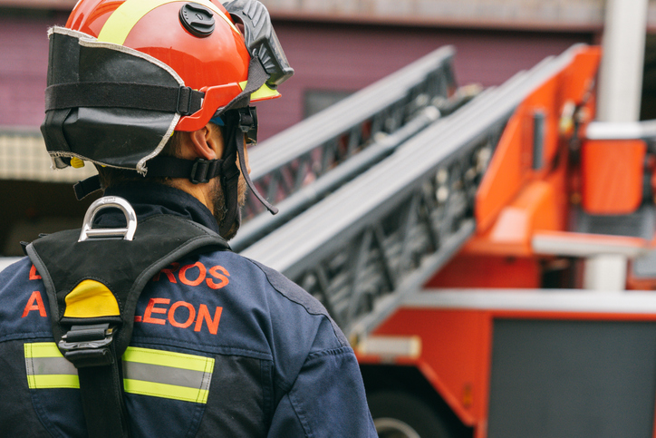 Back view of fireman wearing safety helmet and protective uniform standing near fire engine with ladder