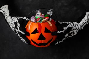 Jack O' Lantern, trick or treat bucket, filled with candies held by a skeleton hands on black background. Halloween celebration concept.