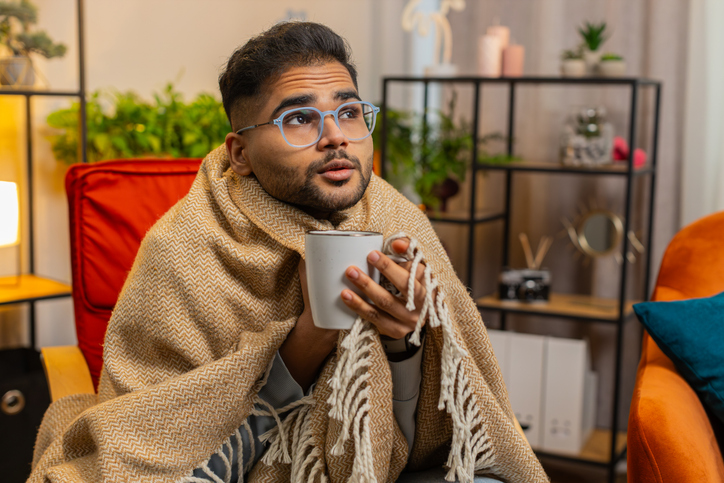 Sick Indian man wrapped in plaid sit alone shivering trembling from cold on chair drinking hot tea