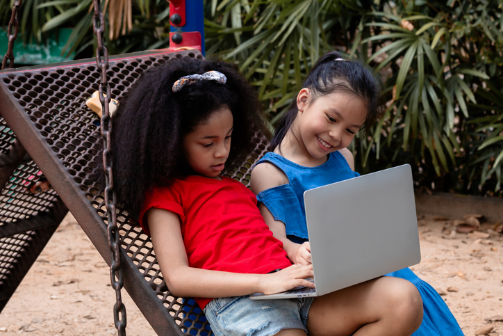 Group of young children multicultural in African-American and Asian girls, spent free time or outing at the playground using laptop. combining outdoor play with technology modern learning or playing