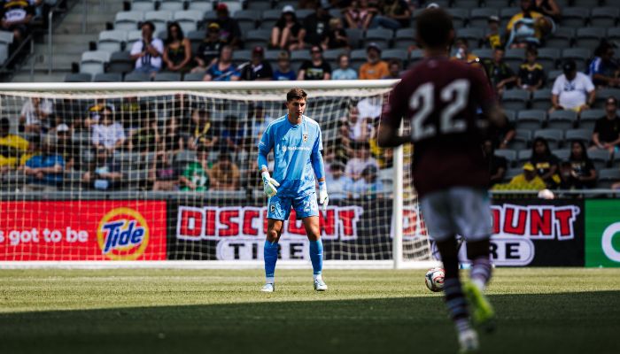 A soccer player in a blue uniform standing in a soccer goal, with a player in a maroon uniform approaching the goal.