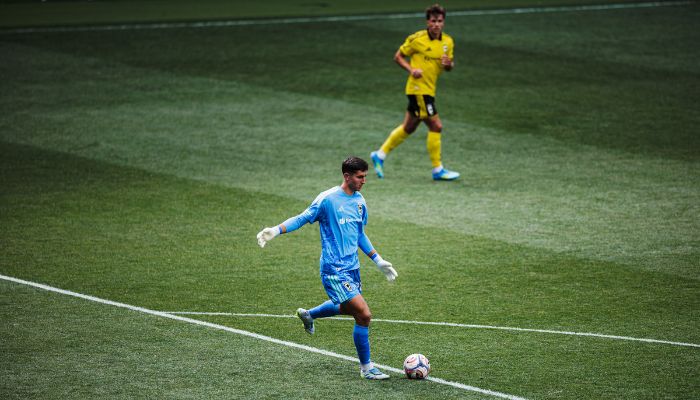 Two soccer players on a field, one in a blue uniform dribbling the ball and the other in a yellow uniform running in the background.