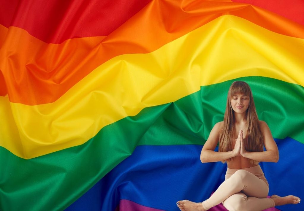 A woman in a yoga pose sitting on the ground in front of a large rainbow flag.