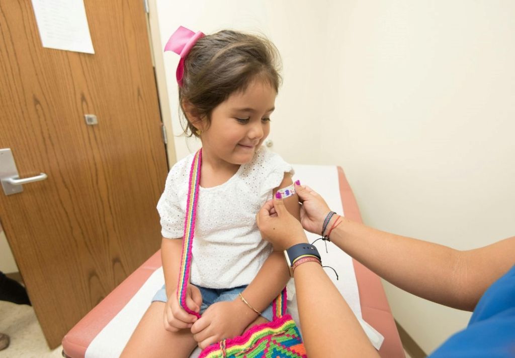 A young girl with a pink bow in her hair sitting in a chair while a medical professional administers a vaccination or injection to her arm.