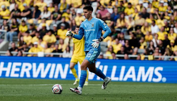 A soccer goalkeeper in a blue uniform running with the ball on a field, with a "Herwin Williams" sign visible in the background.