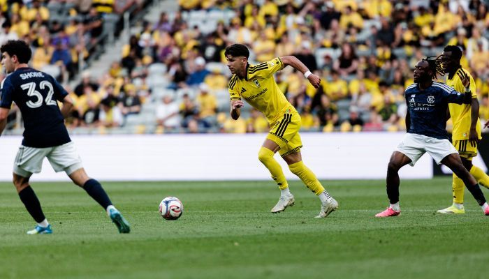 Soccer players in yellow jerseys dribbling the ball on a field, with spectators in the stands.