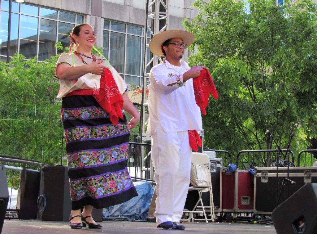 Two people in traditional Mexican clothing performing on a stage surrounded by trees.