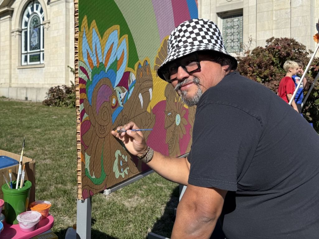 A person wearing a checkered bucket hat and sunglasses painting a colorful mural on an exterior wall.