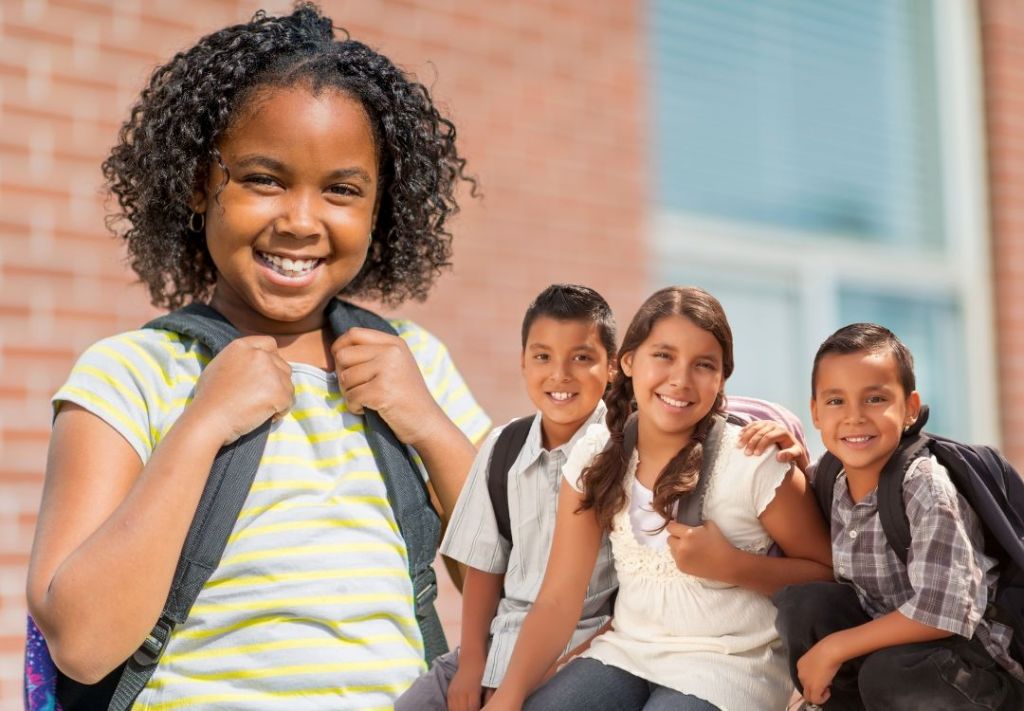 A group of smiling children in school uniforms standing together outdoors in front of a brick building.