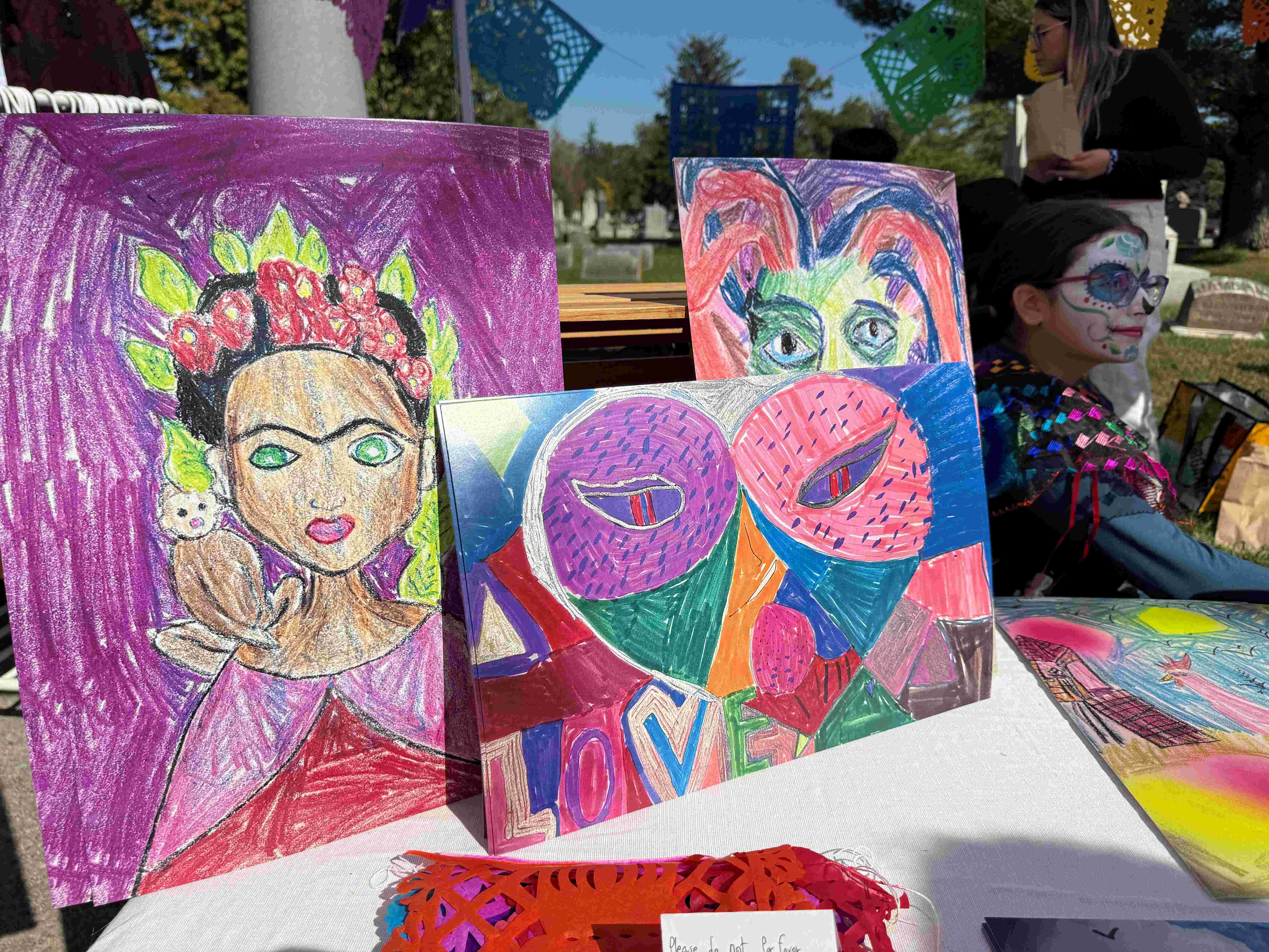Colorful paintings and artwork displayed on a table, including a portrait of a woman with a floral crown and abstract designs.