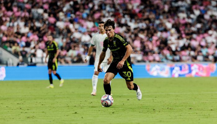 A soccer player in a black jersey dribbling the ball on a soccer field with a large crowd in the background.