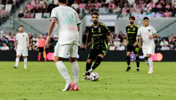 Two soccer players in a match, one in a black jersey dribbling the ball while the other player in a white jersey approaches to defend.