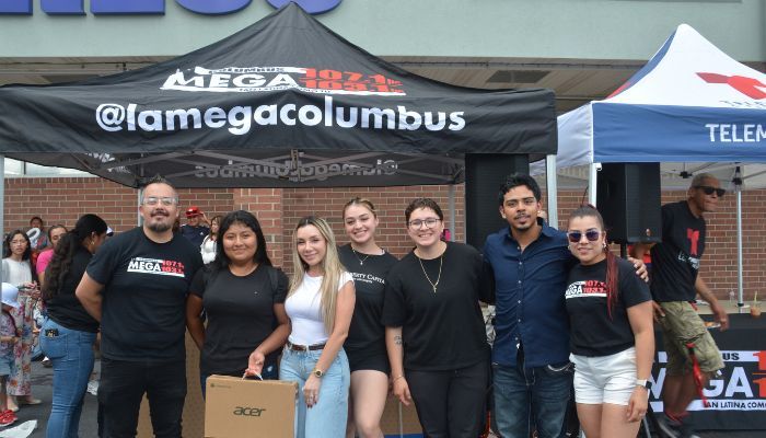 A group of people posing in front of a tent with the MEGA 94.5 logo and "@lamegacolumbus" text.