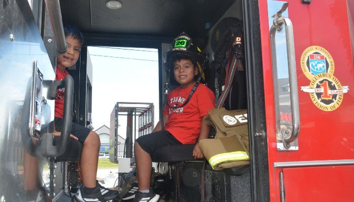 Two young children sitting in the back of a fire engine, wearing firefighter helmets and uniforms.