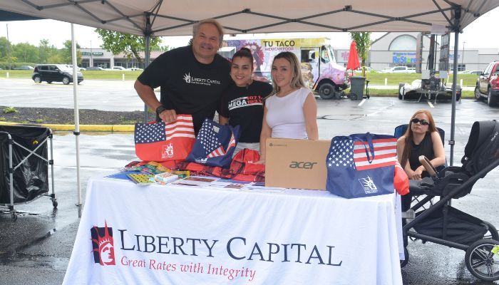 A group of people, including a man and two women, standing behind a table displaying American flags and other patriotic items. The table has the text "Liberty Capital" printed on it.