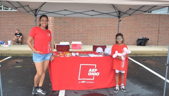 Two people in red shirts standing behind a red table with the AEP Ohio logo, displaying various items on the table.