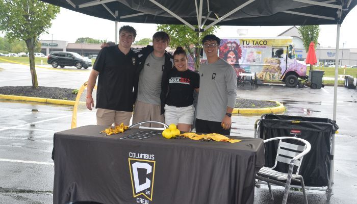 Four young people standing behind a table with a Columbus Crew logo, holding yellow objects in an outdoor setting with a food truck in the background.