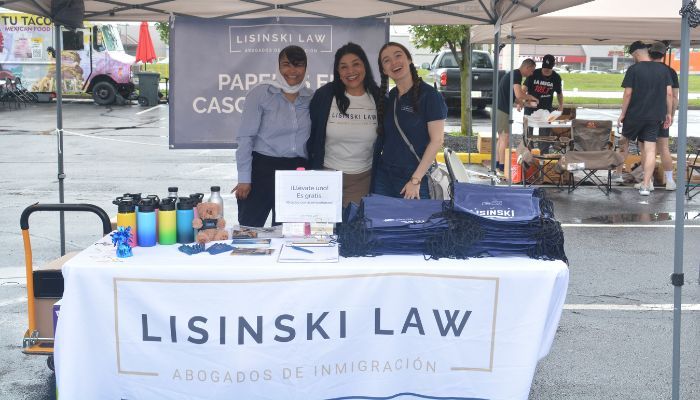 Three young women standing behind a table with the "Lisinski Law" logo, offering information and merchandise at an outdoor event.
