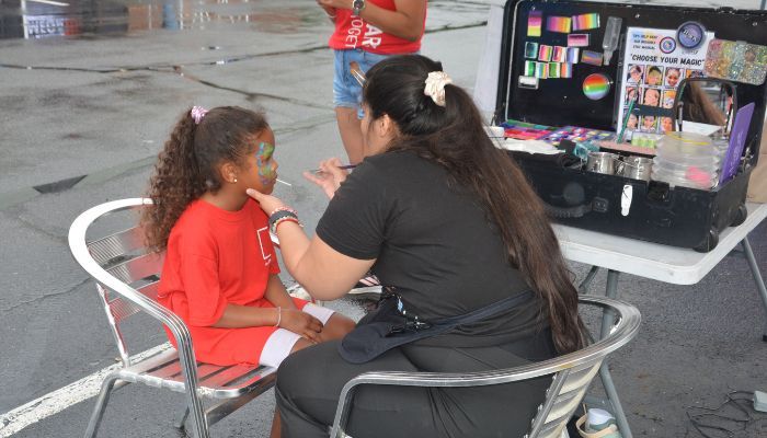 A woman is applying face paint to a young girl's face at an outdoor event or festival.