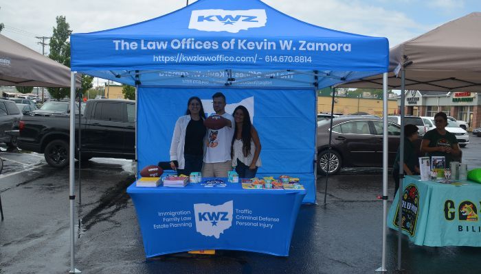 A blue tent with the text "The Law Offices of Kevin W. Zamora" and "KWZ" logo, with three people standing behind a table displaying legal services and materials.