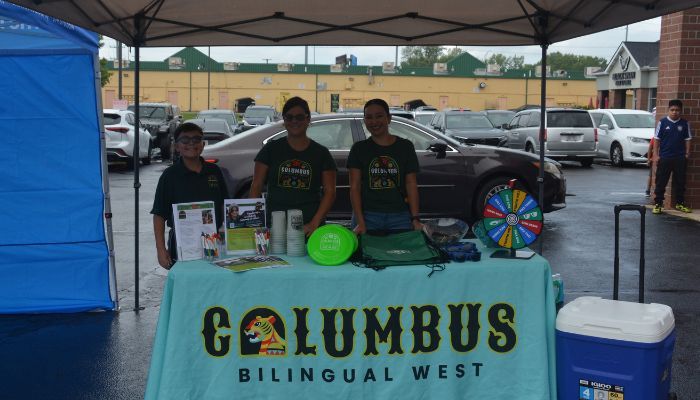 Three people staffing a Calumbus Bilingual West booth at an outdoor event, with a prize wheel and promotional materials on the table.