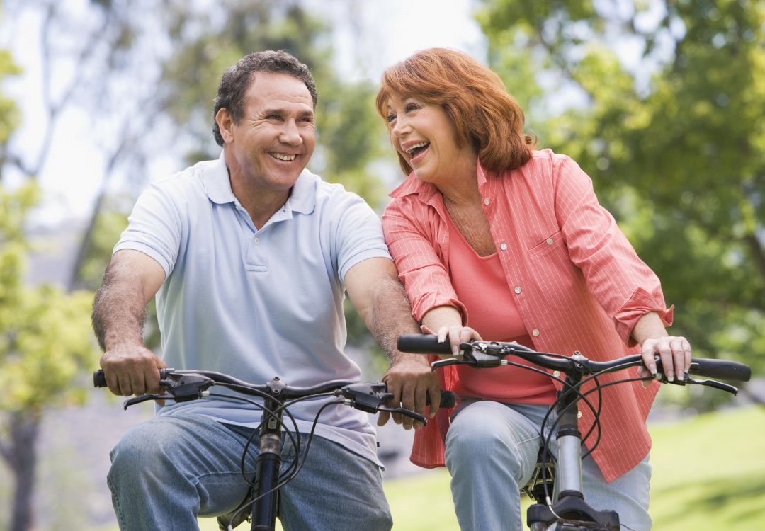 A middle-aged couple riding bicycles and smiling in a park with trees in the background.