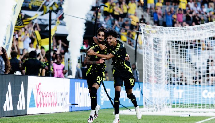 Two soccer players in yellow and black uniforms celebrating a goal on a soccer field with spectators in the background.