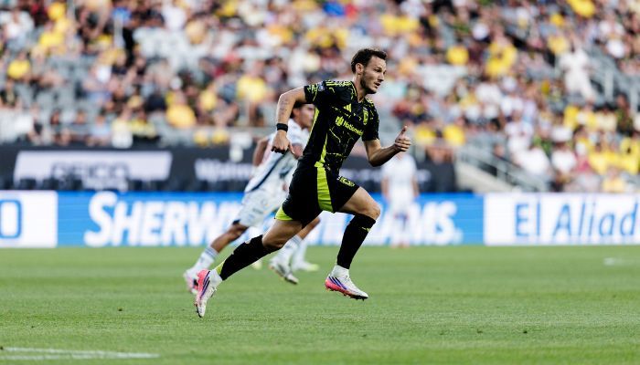 A soccer player in a black and green jersey running on a field with a crowd in the background.