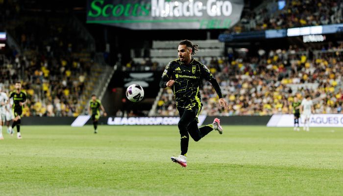 A soccer player in a black uniform running with the ball on a soccer field, with the "Scouts Made Gro" logo visible in the background.
