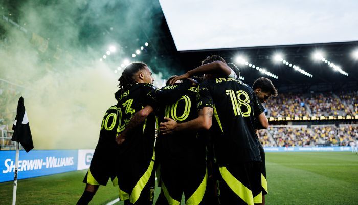 A group of soccer players celebrating together on a field, with smoke and lights in the background.