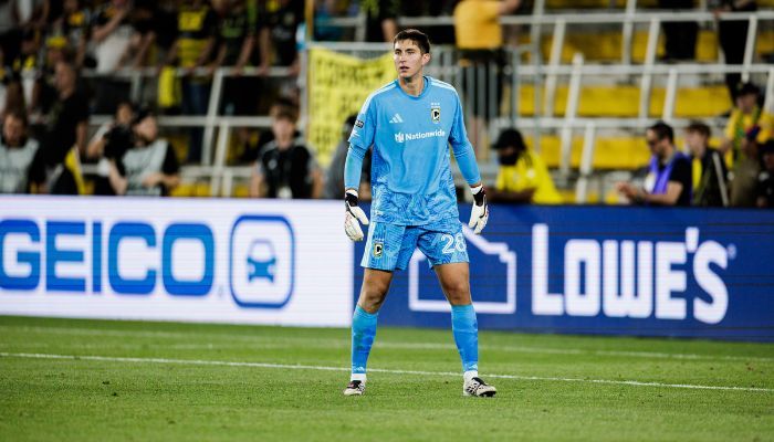 A soccer goalkeeper in a blue uniform stands on a soccer field, with a Geico and Lowe's logo visible in the background.