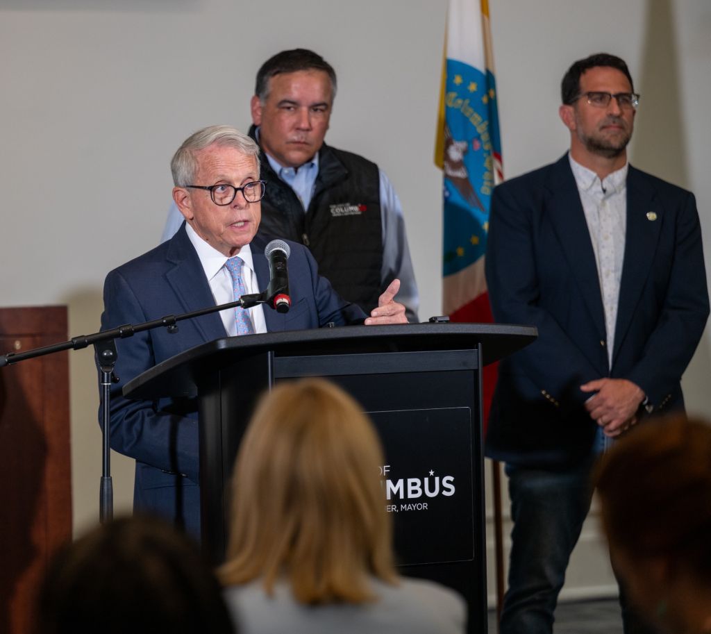 Three men standing at a podium, one speaking into a microphone. The podium has "MAYOR" printed on it.