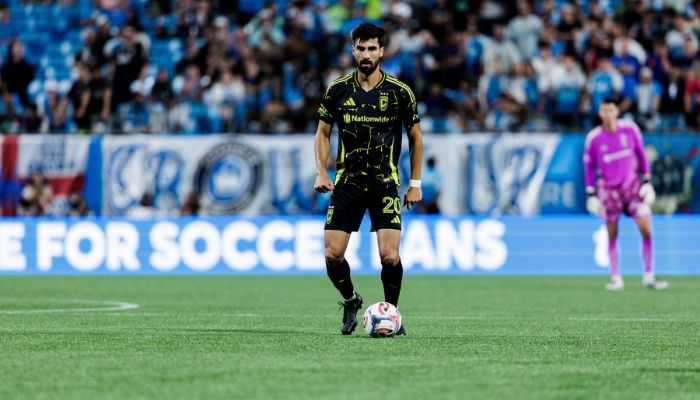 A soccer player in a black jersey with the number 20 dribbling a ball on a soccer field, with a "For Soccer Fans" sign visible in the background.