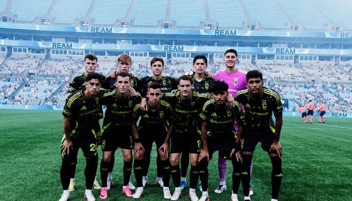 A soccer team posing for a group photo on a stadium field, with the "REAM" logo visible in the background.