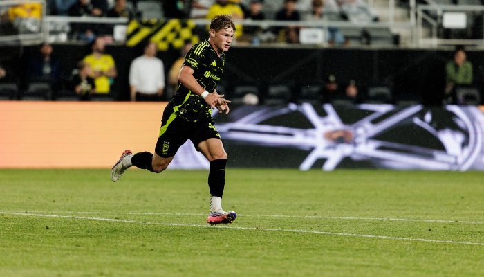A soccer player in a black and yellow uniform running on a field with a large logo visible in the background.