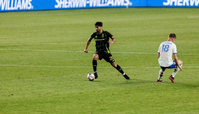 Two soccer players on a field, one in a black jersey dribbling the ball while the other in a white jersey approaches.