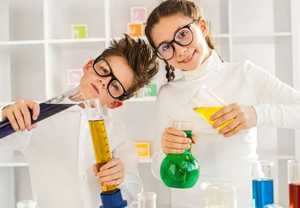 Two young people in lab coats and glasses holding beakers with colored liquids in a science lab.