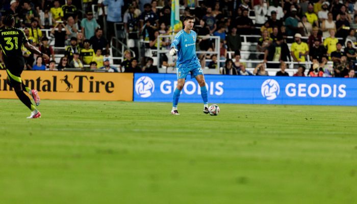 A soccer player in a blue uniform dribbling the ball on a soccer field, with advertising boards visible in the background.