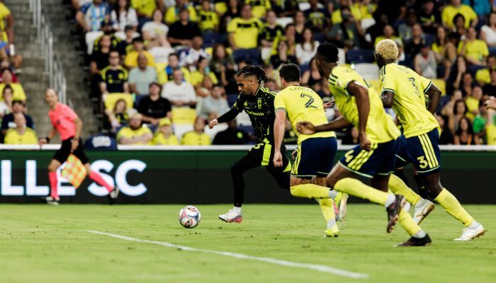 Soccer players in green and yellow jerseys competing on a field, with spectators in the stands.