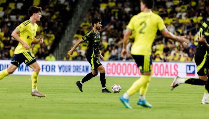 Soccer players in a night match, one player in a black jersey dribbling the ball down the field with "GOOOOOOOOOOALS" displayed on a sign in the background.
