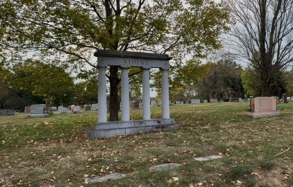 A stone archway with the word "ATHENS" stands in a park with trees and gravestones.